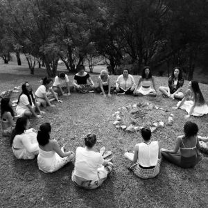 A group of diverse women sits in a circle around a fire pit in a grassy meadow.