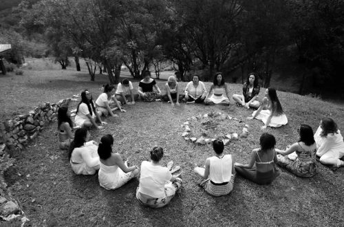 A group of diverse women sits in a circle around a fire pit in a grassy meadow.