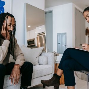 Attentive African American female with clipboard taking notes near sad male partner touching face on couch at home