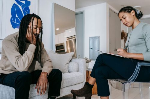 Attentive African American female with clipboard taking notes near sad male partner touching face on couch at home