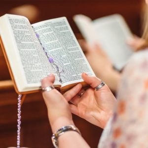 A woman holding a Bible and rosary beads, engaged in prayer inside a church.
