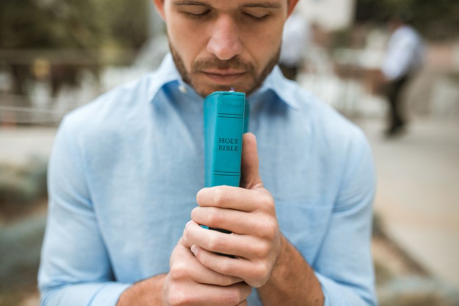 Young man in blue shirt holding a Holy Bible while praying outdoors.