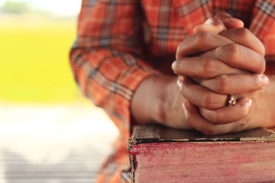 Close-up of clasped hands resting on a book outdoors, symbolizing prayer and reflection.