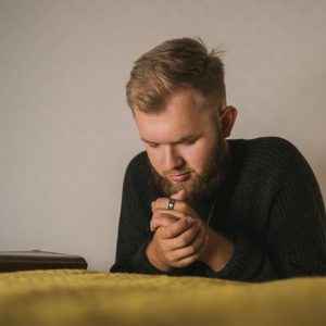 Bearded man in a knitted sweater praying with a Bible beside him in a serene indoor setting.