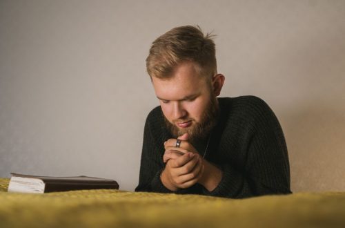 Bearded man in a knitted sweater praying with a Bible beside him in a serene indoor setting.