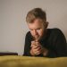 Bearded man in a knitted sweater praying with a Bible beside him in a serene indoor setting.