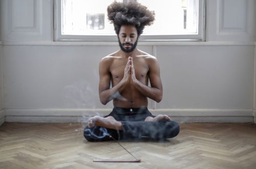 Shirtless man meditating in lotus pose indoors with incense smoke creating a serene atmosphere.