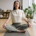 Woman sitting cross-legged on a yoga mat indoors, eyes closed, conveying peace and relaxation.