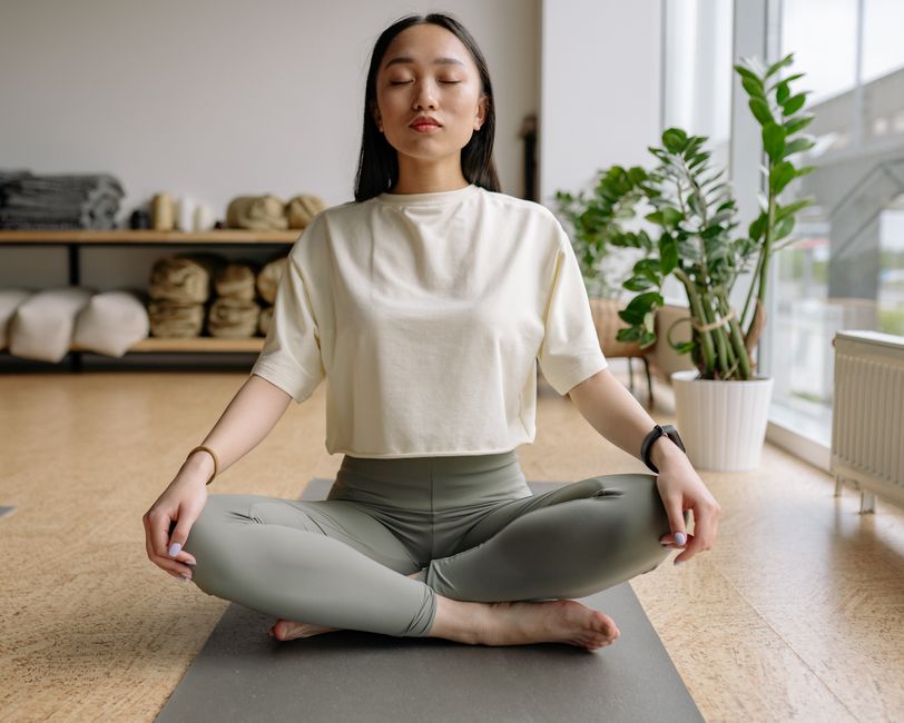 Woman sitting cross-legged on a yoga mat indoors, eyes closed, conveying peace and relaxation.