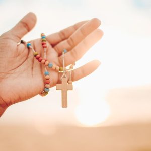 A hand holding a colorful rosary with a wooden cross against a bright sunrise background.