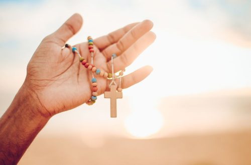 A hand holding a colorful rosary with a wooden cross against a bright sunrise background.