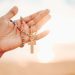 A hand holding a colorful rosary with a wooden cross against a bright sunrise background.