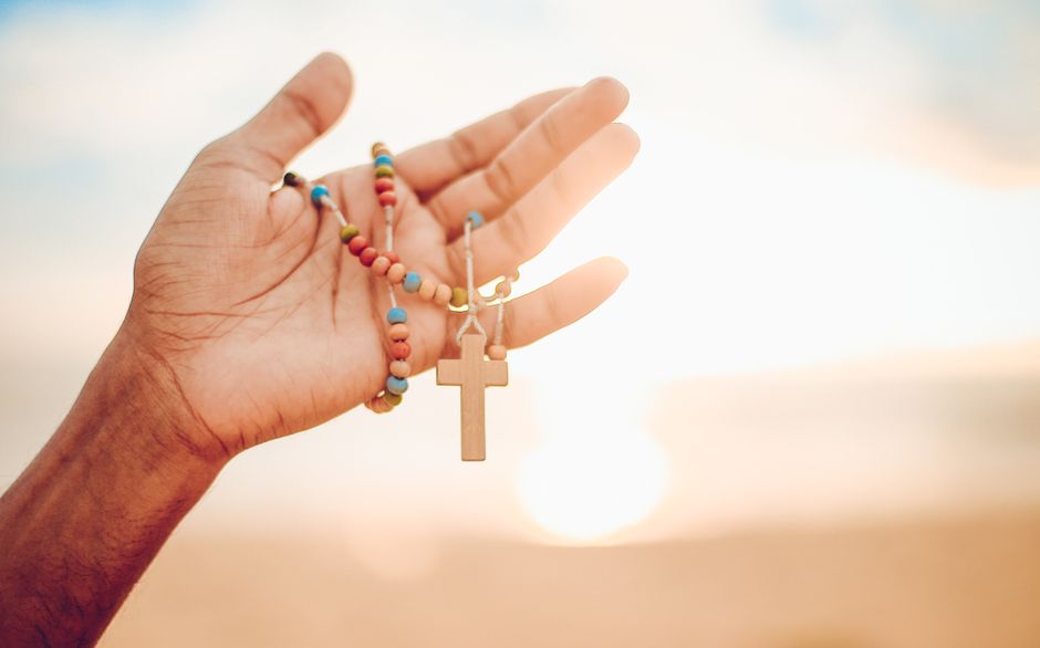 A hand holding a colorful rosary with a wooden cross against a bright sunrise background.