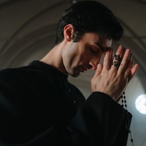 A priest in a black gown solemnly prays with rosary beads in a serene church setting.