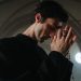 A priest in a black gown solemnly prays with rosary beads in a serene church setting.