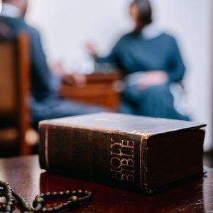 A close-up of a Holy Bible and prayer beads on a wooden table with blurred people in background.