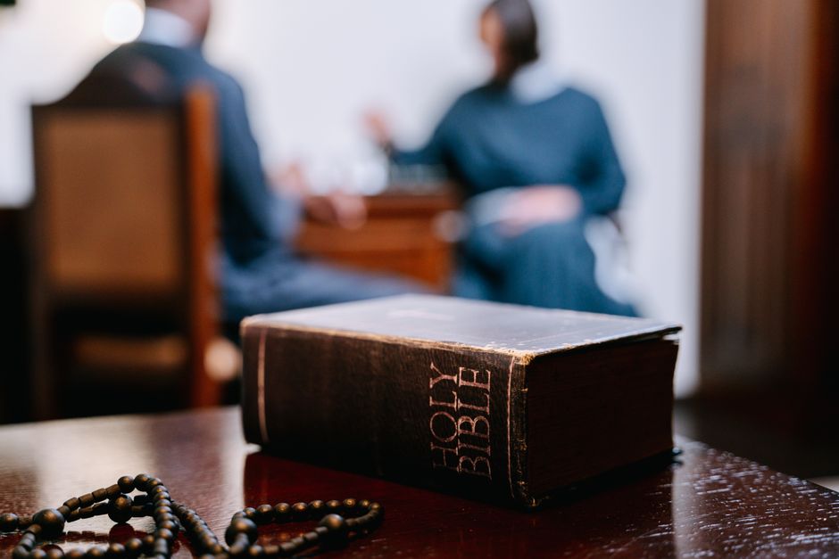 A close-up of a Holy Bible and prayer beads on a wooden table with blurred people in background.