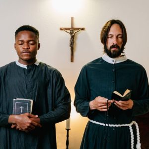 Two priests standing indoors with closed eyes, holding religious items, in a moment of prayer.