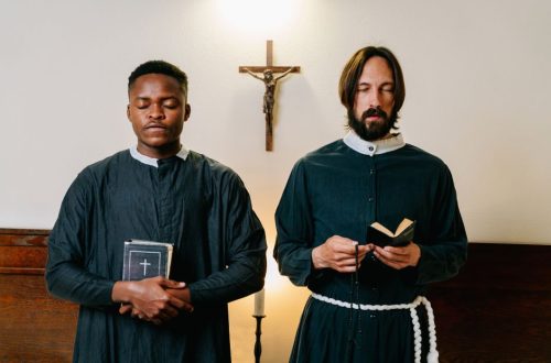 Two priests standing indoors with closed eyes, holding religious items, in a moment of prayer.