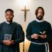 Two priests standing indoors with closed eyes, holding religious items, in a moment of prayer.