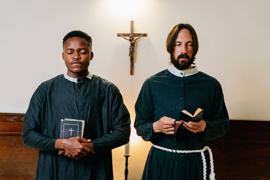 Two priests standing indoors with closed eyes, holding religious items, in a moment of prayer.