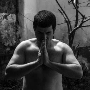 Black and white portrait of a shirtless man meditating, with hands clasped outdoors.