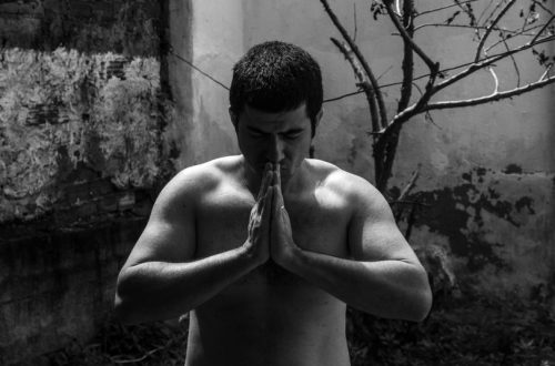Black and white portrait of a shirtless man meditating, with hands clasped outdoors.