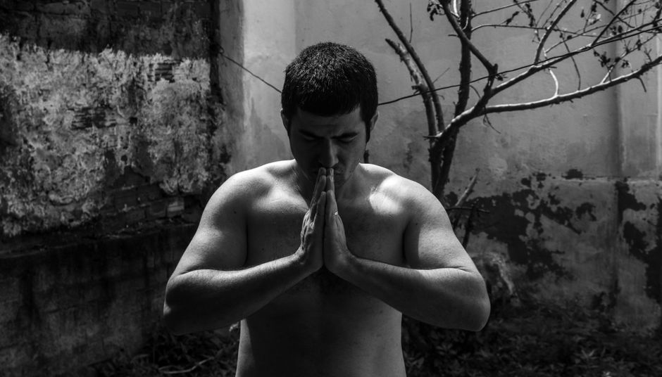 Black and white portrait of a shirtless man meditating, with hands clasped outdoors.