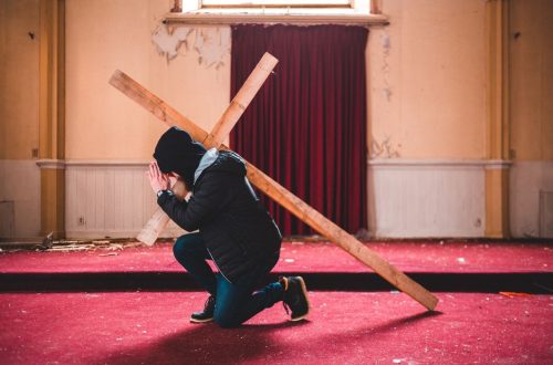 A person kneeling in prayer, carrying a wooden cross inside an abandoned chapel with red carpet.