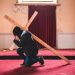 A person kneeling in prayer, carrying a wooden cross inside an abandoned chapel with red carpet.