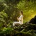 A woman meditating outdoors with prayer beads amidst lush forest greenery.