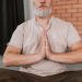 Senior man practicing meditation with hands in prayer pose inside a calm setting.
