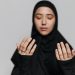A young Muslim woman in traditional attire prays with her hands raised and eyes closed, symbolizing devotion.