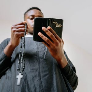 A man in a cassock holds a rosary and reads the Bible indoors, symbolizing faith and devotion.