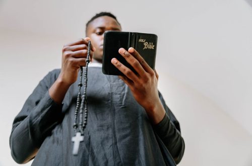 A man in a cassock holds a rosary and reads the Bible indoors, symbolizing faith and devotion.