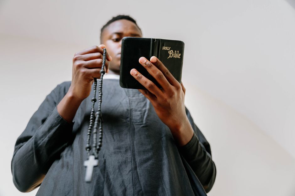 A man in a cassock holds a rosary and reads the Bible indoors, symbolizing faith and devotion.