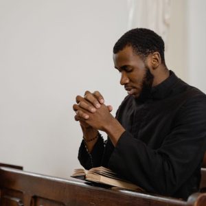 A thoughtful man in a church pew praying with hands clasped and eyes closed.