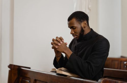 A thoughtful man in a church pew praying with hands clasped and eyes closed.