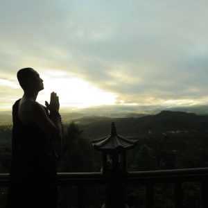 A monk in silhouette prays peacefully at sunrise with mountains in the background.