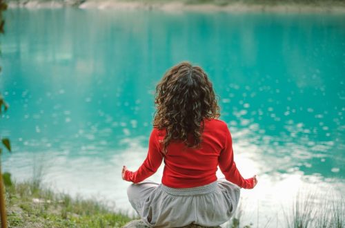 A woman in red meditates by a tranquil blue lake, reflecting calm and serenity.