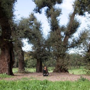 A woman practices meditation among towering trees in a peaceful outdoor setting.