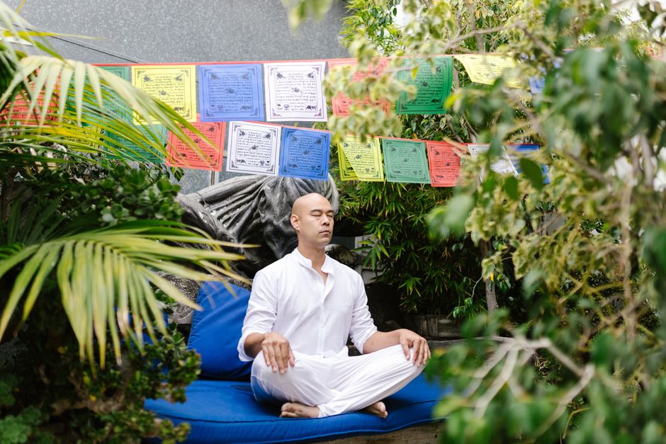 A man in white meditates outdoors in a garden, surrounded by vibrant prayer flags and greenery.