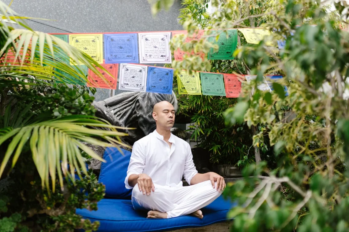 Homem meditando em um jardim, rodeado por bandeirinhas coloridas e vegetação, ideal para um espaço de meditação