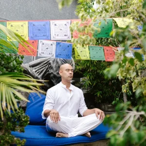 Homem meditando em um jardim, rodeado por bandeirinhas coloridas e vegetação, ideal para um espaço de meditação