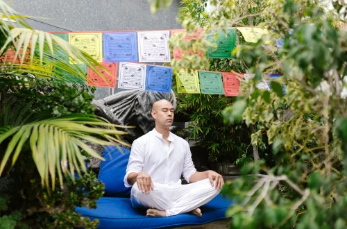 Homem meditando em um jardim, rodeado por bandeirinhas coloridas e vegetação, ideal para um espaço de meditação