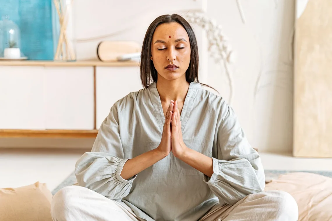 Mulher meditando com os olhos fechados e as mãos unidas em um ambiente interno tranquilo, simbolizando gratidão na meditação