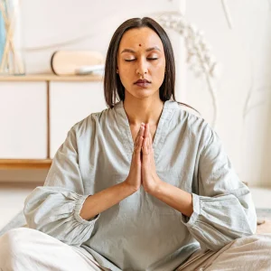 Mulher meditando com os olhos fechados e as mãos unidas em um ambiente interno tranquilo, simbolizando gratidão na meditação